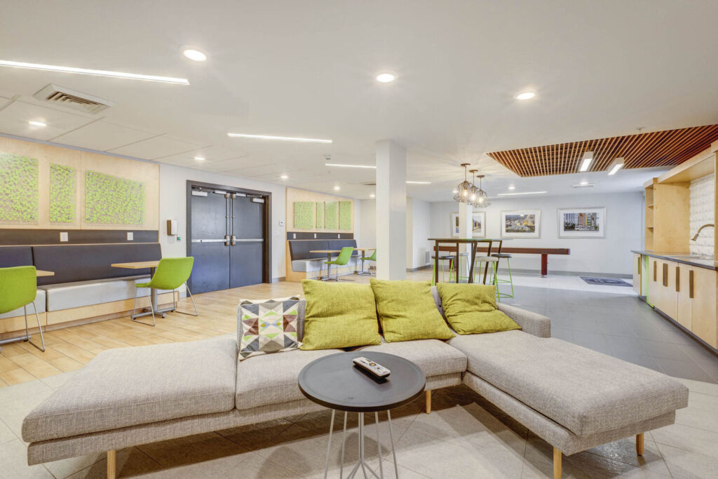 The inside of the clubhouse featuring a grey couches and green chairs with yellow accent pillows for residents to relax and socialize.